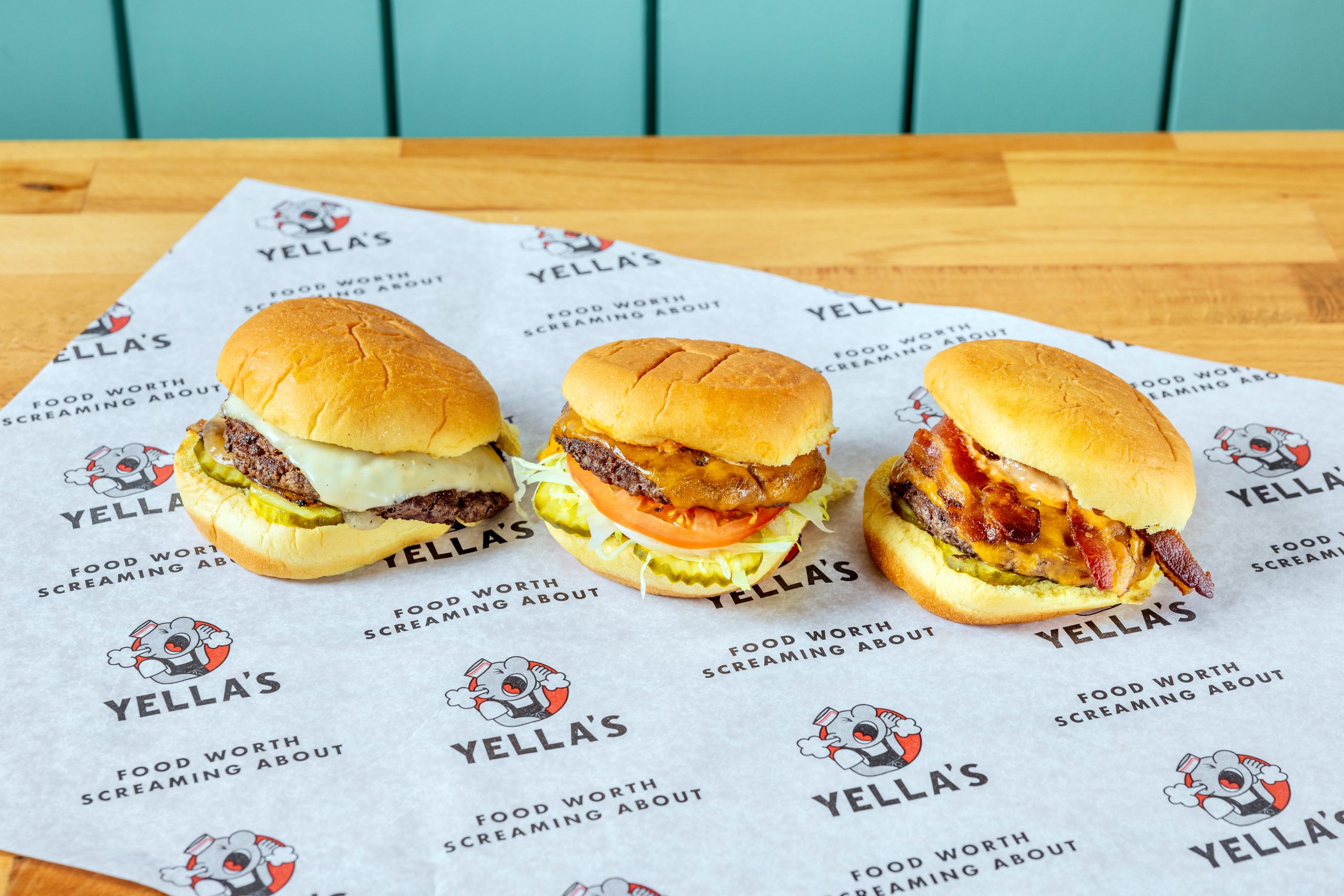 An overhead view of various sandwiches served with waffle fries and crispy onion rings on branded Yella's paper.