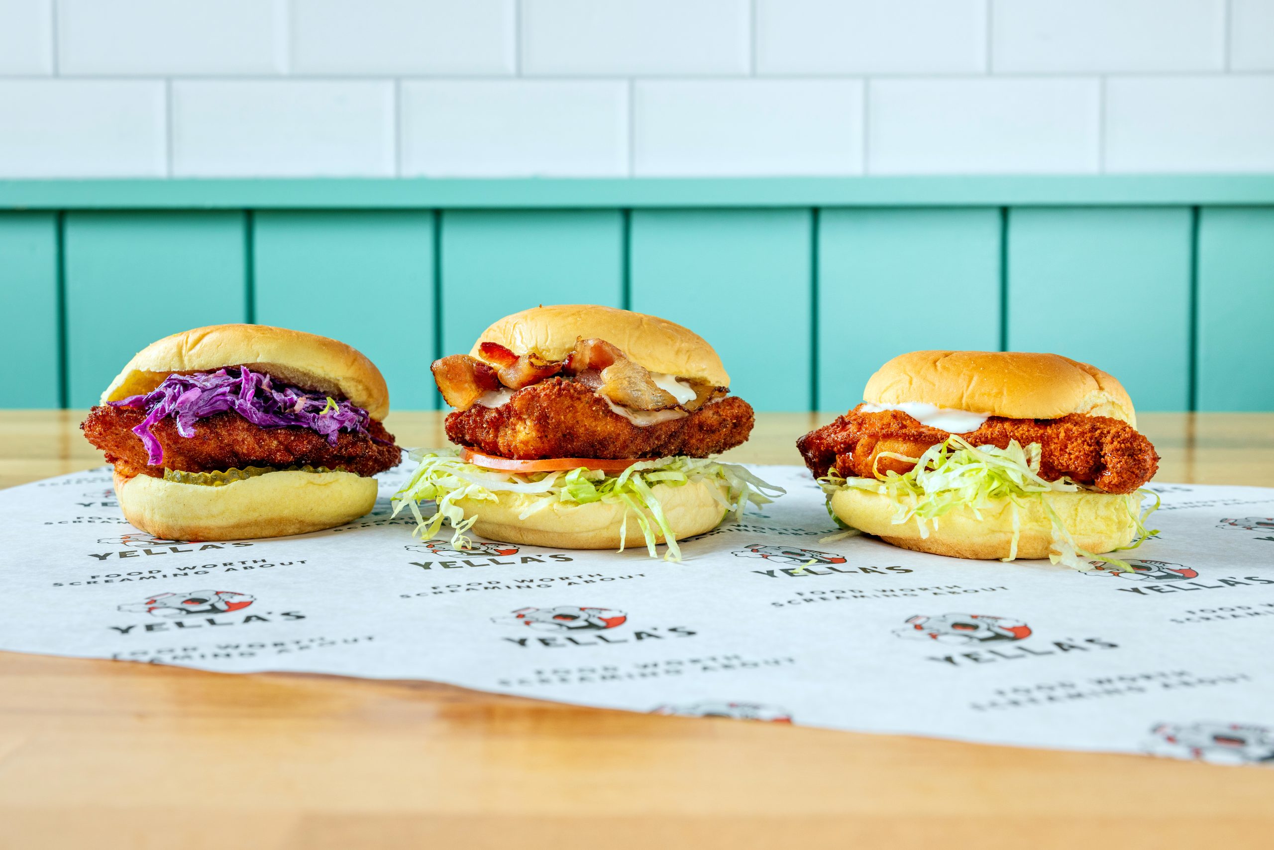 Three Yella's chicken sandwiches on branded paper: one with purple slaw, one with bacon and lettuce, and one with shredded lettuce. All feature breaded chicken on golden buns, displayed on a wooden table against teal paneling. The paper shows Yella's logo and tagline