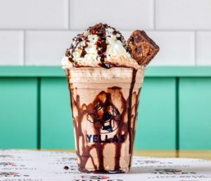 A chocolate milkshake in a Yella's branded cup with chocolate drizzle, whipped cream, chocolate sprinkles, and a brownie piece on top, photographed against teal paneling.