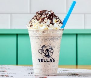 A cookies and cream milkshake in a Yella's branded cup with whipped cream, chocolate drizzle, cookie crumbles, and a blue straw, photographed against teal paneling.