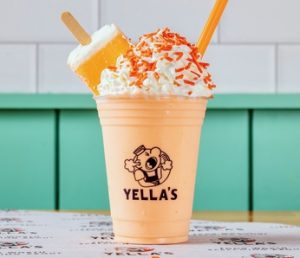 An orange creamsicle milkshake in a Yella's branded cup with whipped cream, orange sprinkles, and an orange straw, photographed against teal paneling