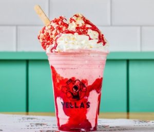 A pink milkshake in a Yella's branded cup made with strawberry shortcake ice cream bar pieces, topped with whipped cream, red drizzle, and a beige straw, photographed against teal paneling