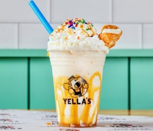 A vanilla milkshake in a Yella's branded cup with yellow drizzle, whipped cream, rainbow sprinkles, a cookie piece, and a blue straw, photographed against teal paneling.